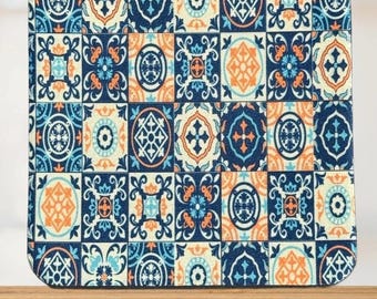 May include: Square coaster with a repeating pattern of ornate, geometric tiles in shades of navy blue, orange, and light blue. The design features floral and cross-like motifs, creating a decorative and artistic look. The coaster has rounded corners.