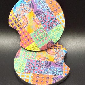 May include: Two round car coasters with a colorful patchwork design. The coasters feature a mix of patterns in shades of blue, purple, orange, yellow, and green. Each coaster has a curved cutout for easy removal from a cup holder.