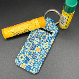 May include: A blue and orange patterned lip balm holder with a silver keyring, holding a yellow lip balm tube. Another green and yellow lip balm tube is also present. The holder has a geometric tile design.