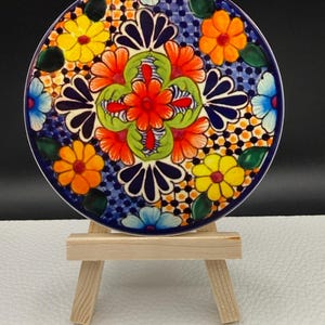 May include: A round, hand-painted ceramic plate with a vibrant floral design. The plate features a dark blue border and a central pattern of orange, yellow, and blue flowers. The plate is displayed on a small wooden easel.