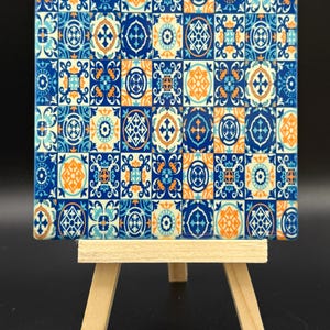 May include: Square ceramic tile with a repeating pattern of blue and orange geometric designs. The tile is displayed on a small wooden easel against a black background. The tile's design is reminiscent of traditional Portuguese tiles.