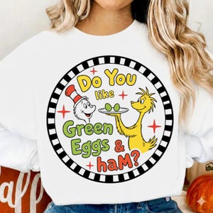 Op de afbeelding: Witte sweatshirt met een cirkelvormige afbeelding. De afbeelding toont de tekst "Do You like Green Eggs & ham?" in geel en groen, samen met stripfiguren en een zwart-witte rand.