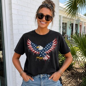 May include: Black t-shirt featuring a patriotic design of an eagle with outstretched wings. The eagle's wings are designed with the American flag's stars and stripes. The shirt is worn with blue jeans and sunglasses.