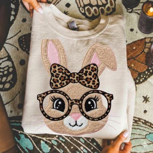 Pode incluir: Camisola bege com um bordado de rosto de coelho com óculos e laço. O coelho tem orelhas grandes com interior rosa, óculos redondos e laço com estampa de leopardo. A camisola está dobrada e é segurada por mãos.