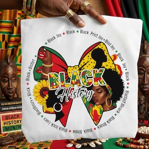 May include: White tote bag with a vibrant bow design, displaying the words "BLACK HISTORY". The bow incorporates images of Black women, sunflowers, and the text "Black Joy" and "Black". The bag is held by a person with gold rings.