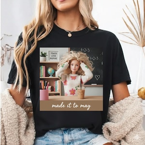 Peut inclure: T-shirt noir avec un graphisme représentant une poupée dans une salle de classe. La poupée porte un pull à fleurs devant un tableau noir avec des équations mathématiques. Le texte "made it to may" est en bas du graphisme.
