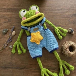 May include: A crocheted frog doll in green and blue. The frog is wearing blue dungarees with a yellow star. It has large eyes, a wide mouth, and long limbs. The doll is lying on a wooden surface. The frog is a handmade item.