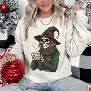 May include: Cream-colored sweatshirt featuring a skeleton wearing a witch hat, scarf, and sweater, holding a steaming mug. The witch hat has a poinsettia and crescent moon. The scarf is green and red plaid. A Christmas tree and ornament are in the background.
