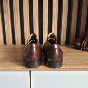 May include: A pair of brown leather dress shoes, viewed from the back, sit on a wooden shelf. The shoes have a polished finish and a classic design. The background features a wooden slat wall.