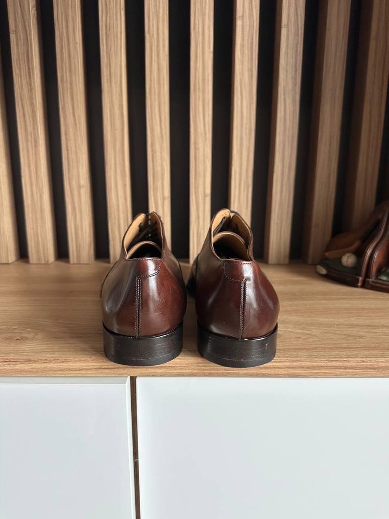 May include: A pair of brown leather dress shoes, viewed from the back, sit on a wooden shelf. The shoes have a polished finish and a classic design. The background features a wooden slat wall.