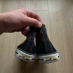 Puede incluir: Zapatillas altas negras con suelas blancas y un emblema decorativo con un ojo alado. Las zapatillas tienen una parte superior negra con detalles dorados y marrones y costuras blancas.