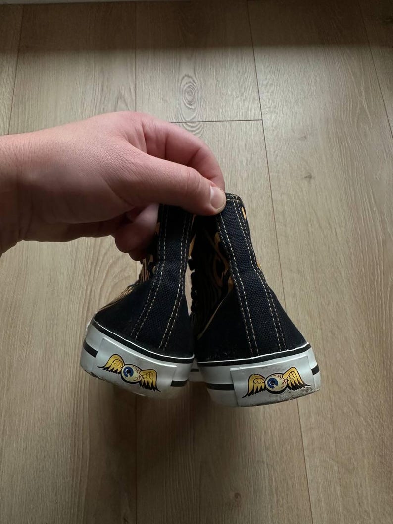 Puede incluir: Zapatillas altas negras con suelas blancas y un emblema decorativo con un ojo alado. Las zapatillas tienen una parte superior negra con detalles dorados y marrones y costuras blancas.