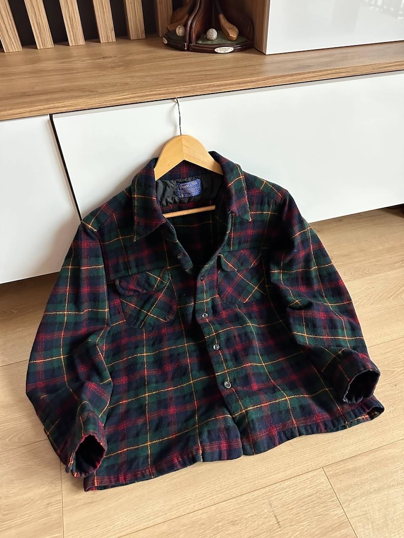 May include: A plaid flannel shirt in a classic pattern of navy, green, red, and yellow. The shirt has two chest pockets, a button-down front, and long sleeves. The shirt is hanging on a wooden hanger.