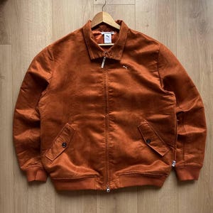 May include: A burnt orange jacket with a collared neckline, a zipper closure, and two front pockets. The jacket features the Puma logo on the chest and is made of a soft, textured material. The jacket is hanging on a wooden hanger.