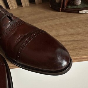 May include: Close-up of a brown leather dress shoe with a cap toe and decorative stitching. The shoe has a polished finish and is laced up. The image suggests a focus on classic footwear.