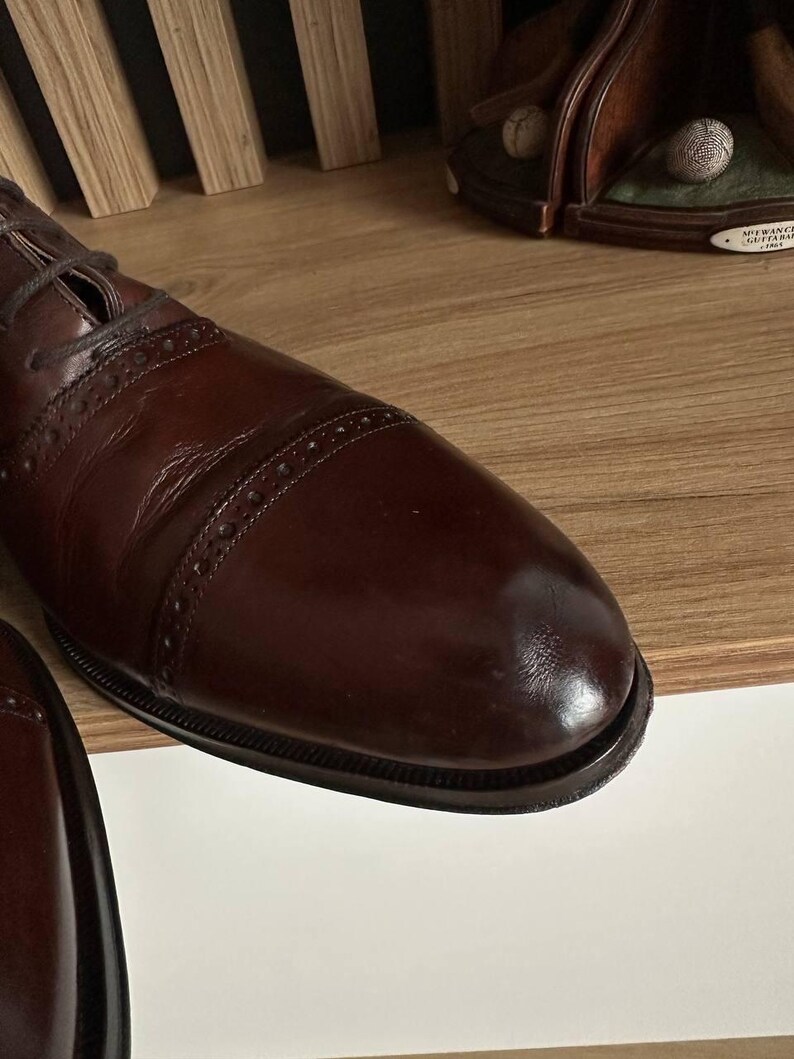 May include: Close-up of a brown leather dress shoe with a cap toe and decorative stitching. The shoe has a polished finish and is laced up. The image suggests a focus on classic footwear.