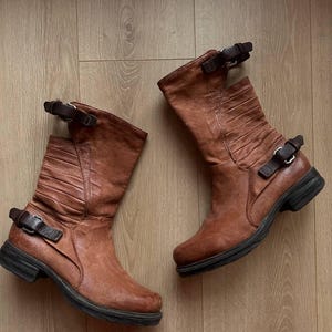May include: A pair of brown leather mid-calf boots with a distressed finish. The boots feature a ridged design on the upper shaft, black soles, and adjustable buckle straps. The boots are displayed on a light brown wooden surface.