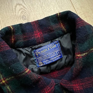 May include: Close-up of a Pendleton wool jacket collar. The jacket features a classic plaid pattern in red, green, and navy blue. A blue label with white text is sewn into the collar, indicating the brand and material.