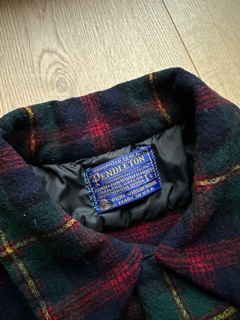 May include: Close-up of a Pendleton wool jacket collar. The jacket features a classic plaid pattern in red, green, and navy blue. A blue label with white text is sewn into the collar, indicating the brand and material.