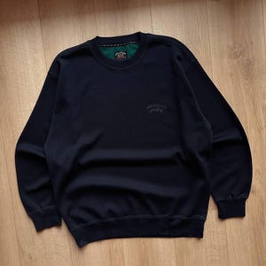 May include: Navy blue sweatshirt with a crew neck and long sleeves. The sweatshirt has a small logo on the chest and a green and black label on the inside of the collar. The fabric appears to be a soft, comfortable material.