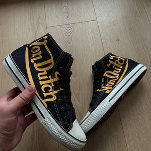 Puede incluir: Zapatillas altas negras con la inscripci&oacute;n dorada "Von Dutch". Las zapatillas tienen suelas y punteras blancas y cordones negros. Los zapatos son de lona.
