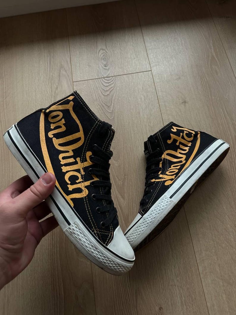 Puede incluir: Zapatillas altas negras con la inscripci&oacute;n dorada "Von Dutch". Las zapatillas tienen suelas y punteras blancas y cordones negros. Los zapatos son de lona.