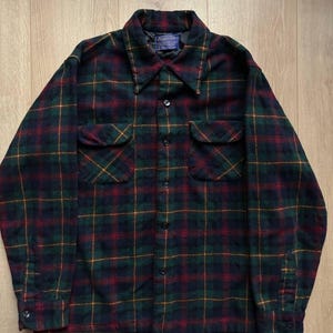 May include: A dark green, red, and yellow plaid flannel shirt with two chest pockets and a button-down front. The shirt has a collar and long sleeves with buttoned cuffs. A small tag is visible near the collar.