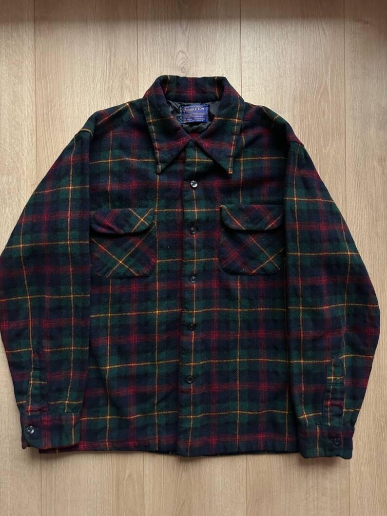 May include: A dark green, red, and yellow plaid flannel shirt with two chest pockets and a button-down front. The shirt has a collar and long sleeves with buttoned cuffs. A small tag is visible near the collar.