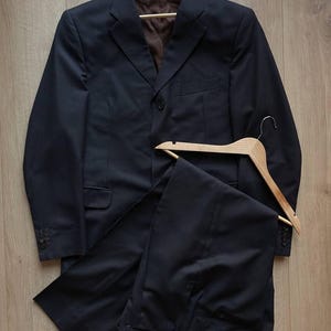 May include: A navy blue suit with subtle pinstripes, consisting of a blazer and trousers, displayed on wooden hangers. The blazer features a two-button closure and a brown interior lining. The suit is set against a light brown wooden background.