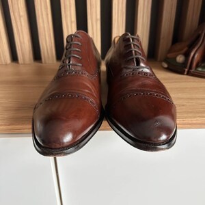 May include: A pair of brown leather Oxford shoes with intricate brogue detailing. The shoes have a classic design with a rounded toe and a lace-up closure. The leather has a polished finish, and the soles are dark.