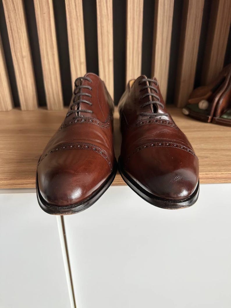 May include: A pair of brown leather Oxford shoes with intricate brogue detailing. The shoes have a classic design with a rounded toe and a lace-up closure. The leather has a polished finish, and the soles are dark.