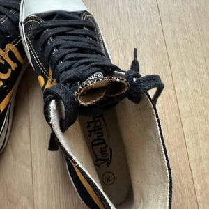 Puede incluir: Zapatillas altas negras y amarillas con punteras blancas y cordones negros. Las zapatillas tienen el logo "Von Dutch" en el interior. Los zapatos son de lona y tienen suela de goma.