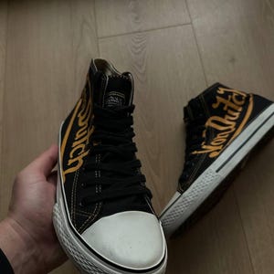 Puede incluir: Zapatillas altas negras con suelas blancas y puntera blanca. Las zapatillas presentan la inscripci&oacute;n "Von Dutch" en amarillo en los laterales. Los zapatos tienen cordones negros y una leng&uuml;eta negra.