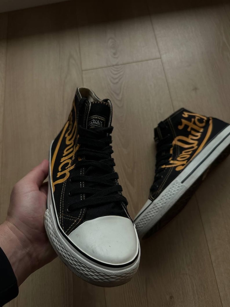 Puede incluir: Zapatillas altas negras con suelas blancas y puntera blanca. Las zapatillas presentan la inscripci&oacute;n "Von Dutch" en amarillo en los laterales. Los zapatos tienen cordones negros y una leng&uuml;eta negra.