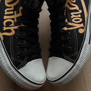 Puede incluir: Par de zapatillas altas negras con punteras y suelas blancas. Las zapatillas presentan la inscripci&oacute;n "Von Dutch" en letras doradas en los laterales. Cordones y costuras negros visibles.