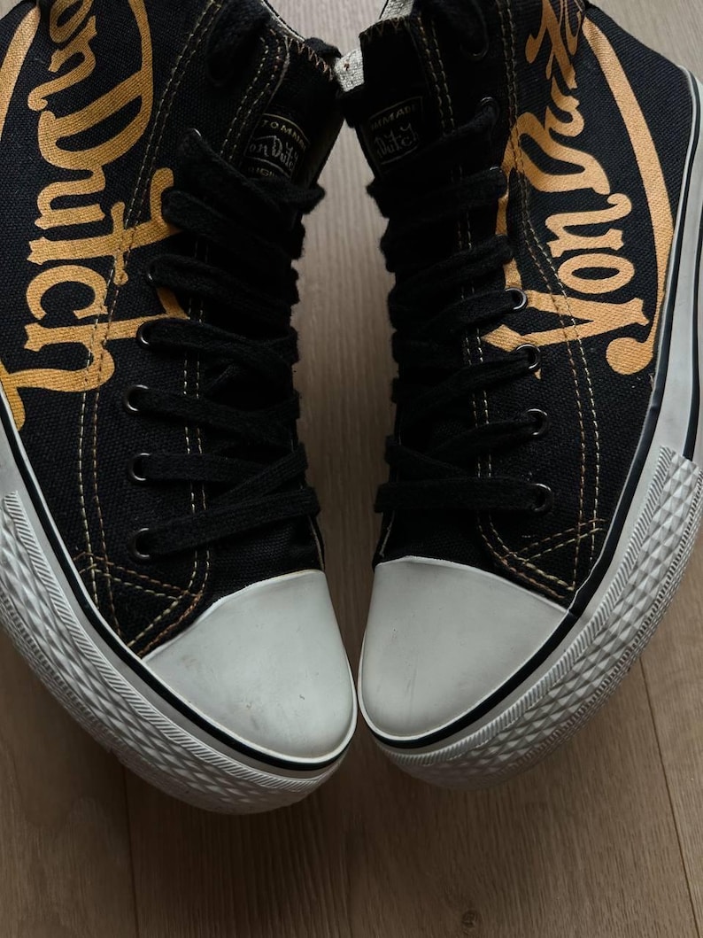 Puede incluir: Par de zapatillas altas negras con punteras y suelas blancas. Las zapatillas presentan la inscripci&oacute;n "Von Dutch" en letras doradas en los laterales. Cordones y costuras negros visibles.