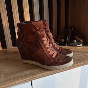 May include: A pair of brown suede wedge sneakers with a side zipper and lace-up closure. The shoes have a white sole and a decorative strap. The background features a wooden shelf and a brown leather golf shoe holder.
