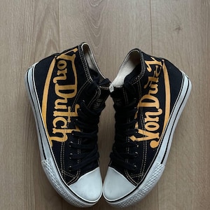 Puede incluir: Un par de zapatillas altas negras con la inscripci&oacute;n "Von Dutch" en letras doradas en los laterales. Las zapatillas tienen suelas y punteras de goma blanca y cordones negros.