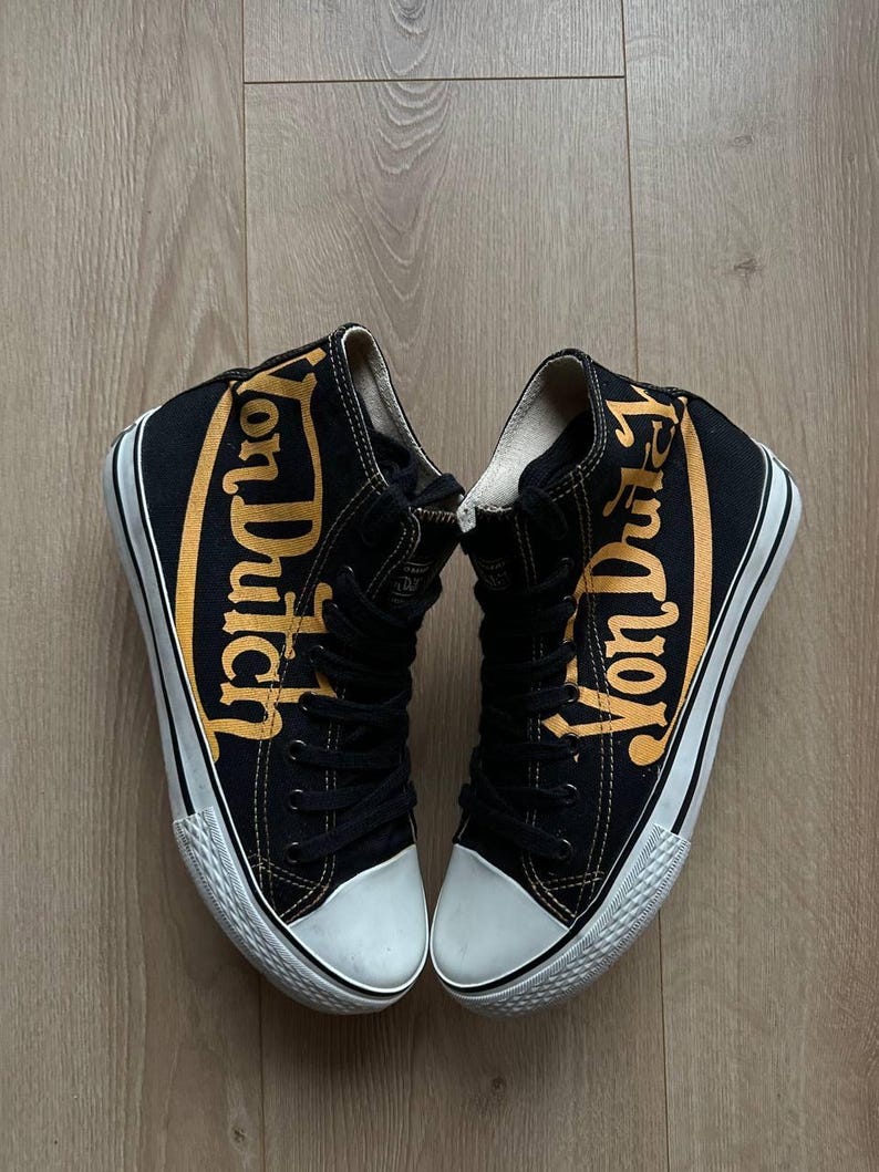 Puede incluir: Un par de zapatillas altas negras con la inscripci&oacute;n "Von Dutch" en letras doradas en los laterales. Las zapatillas tienen suelas y punteras de goma blanca y cordones negros.