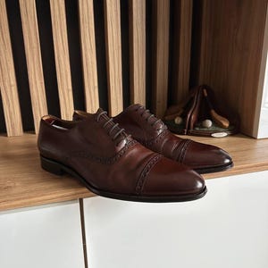 May include: A pair of brown leather dress shoes with cap toes and intricate stitching. The shoes are laced up and sit on a wooden surface. A golf-themed decorative item is visible in the background.