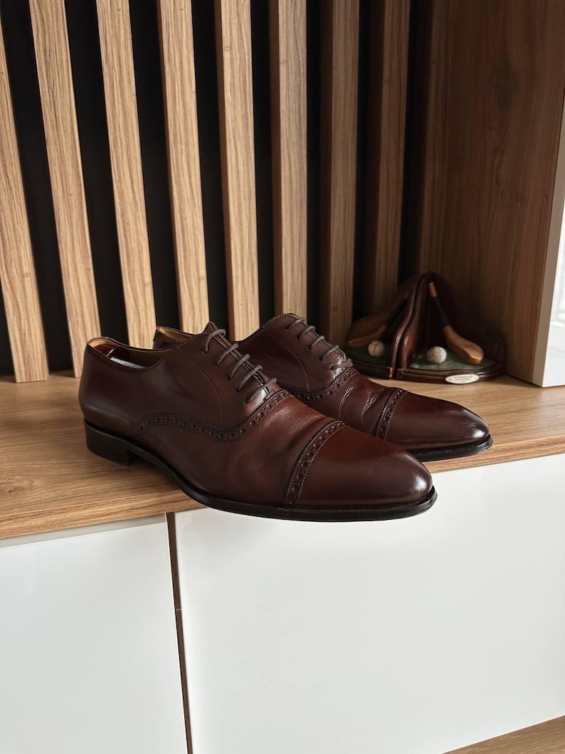 May include: A pair of brown leather dress shoes with cap toes and intricate stitching. The shoes are laced up and sit on a wooden surface. A golf-themed decorative item is visible in the background.