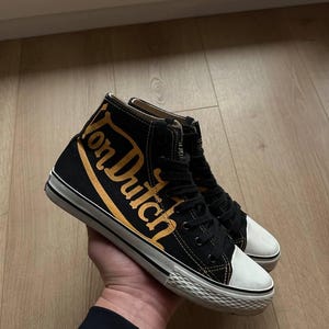 Puede incluir: Zapatillas altas negras con el texto "Von Dutch" en letras doradas. Las zapatillas tienen suelas y punteras de goma blanca. Los zapatos est&aacute;n atados con cordones negros.