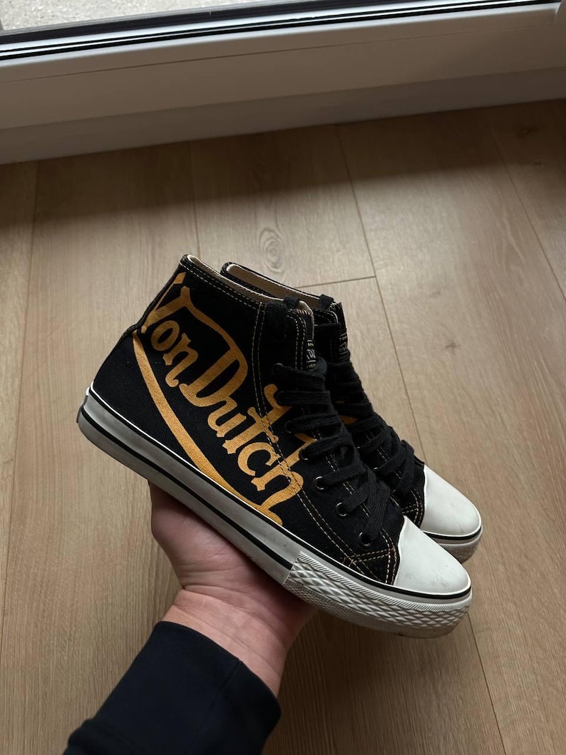 Puede incluir: Zapatillas altas negras con el texto "Von Dutch" en letras doradas. Las zapatillas tienen suelas y punteras de goma blanca. Los zapatos est&aacute;n atados con cordones negros.