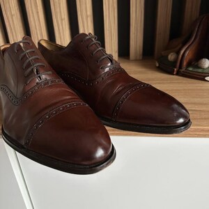 May include: A pair of dark brown leather dress shoes with intricate stitching and lace-up closures. The shoes have a classic design with a polished finish, suitable for formal occasions. The shoes are displayed on a white surface.