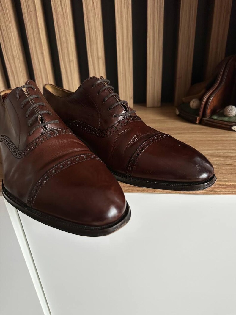 May include: A pair of dark brown leather dress shoes with intricate stitching and lace-up closures. The shoes have a classic design with a polished finish, suitable for formal occasions. The shoes are displayed on a white surface.