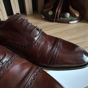 May include: Close-up of two brown leather dress shoes with dark laces. The shoes have a cap-toe design and decorative perforations along the seams. A golf-themed decorative item is in the background.