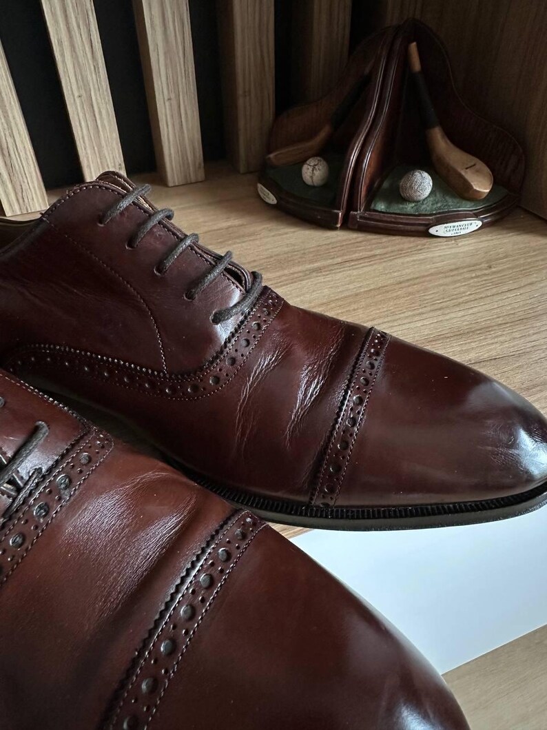 May include: Close-up of two brown leather dress shoes with dark laces. The shoes have a cap-toe design and decorative perforations along the seams. A golf-themed decorative item is in the background.
