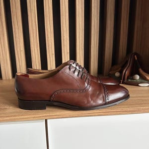May include: A pair of brown leather Oxford shoes with a cap toe and decorative perforations. The shoes have a classic lace-up design and are displayed on a wooden shelf. The shoes are a rich, dark brown color.