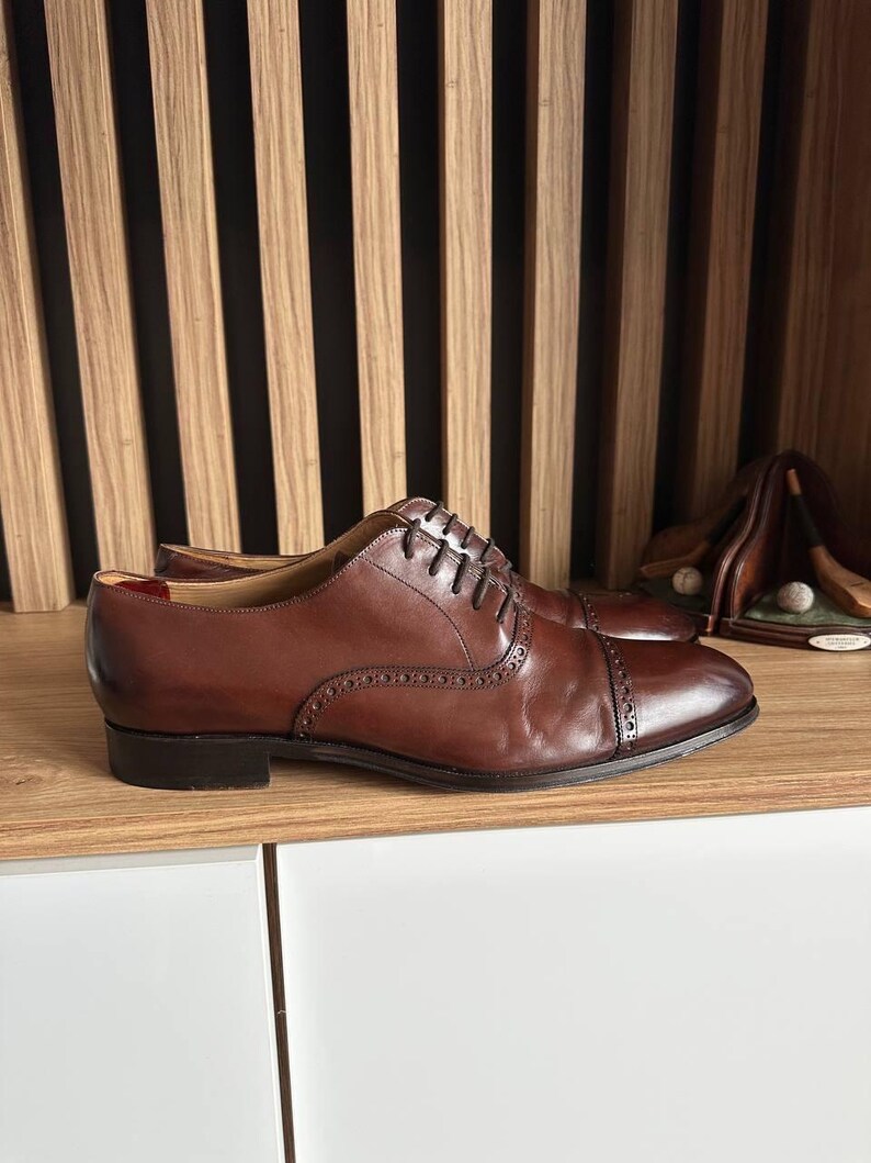 May include: A pair of brown leather Oxford shoes with a cap toe and decorative perforations. The shoes have a classic lace-up design and are displayed on a wooden shelf. The shoes are a rich, dark brown color.