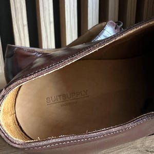 May include: Close-up of a pair of brown leather dress shoes. The interior of the shoe is visible, with the brand name "SUITSUPPLY" and "MADE IN ITALY" stamped on the insole. The shoes have a classic design and are made of high-quality leather.